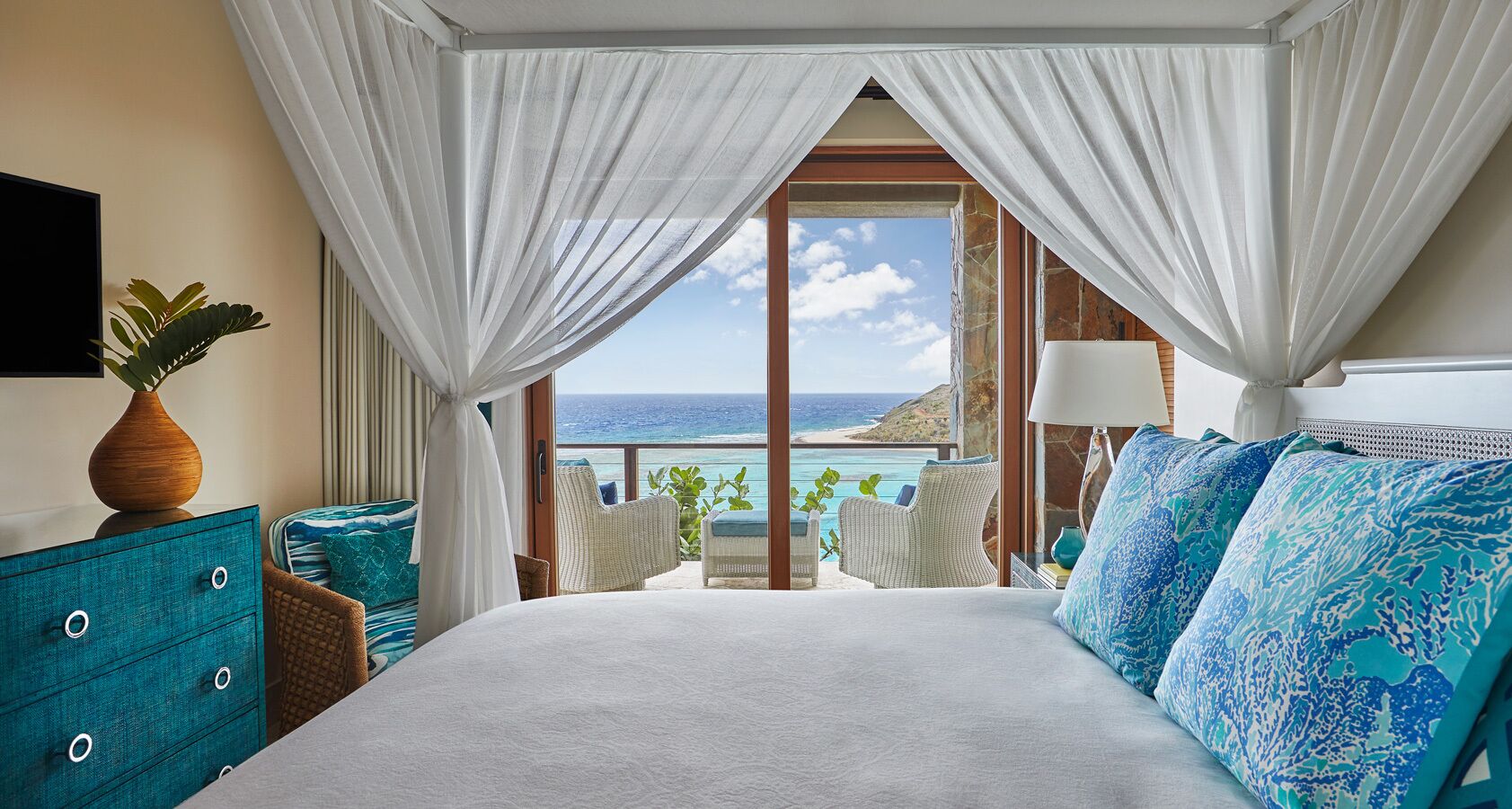 A bedroom with a large bed, a view of the ocean, a balcony with chairs, and a TV on the wall.