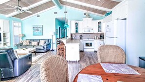 A spacious living area with a high ceiling, a dining table with chairs, a sofa, and a kitchen area with white cabinets and appliances.