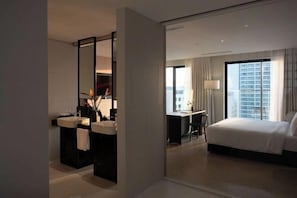 Habitación de hotel con cama, escritorio, silla, ventana grande y baño con espejo y lavamanos.