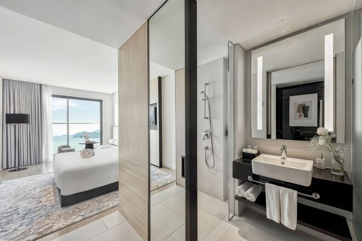 A modern hotel room with a large bed, a bathroom with a vanity, and a view of the sea.