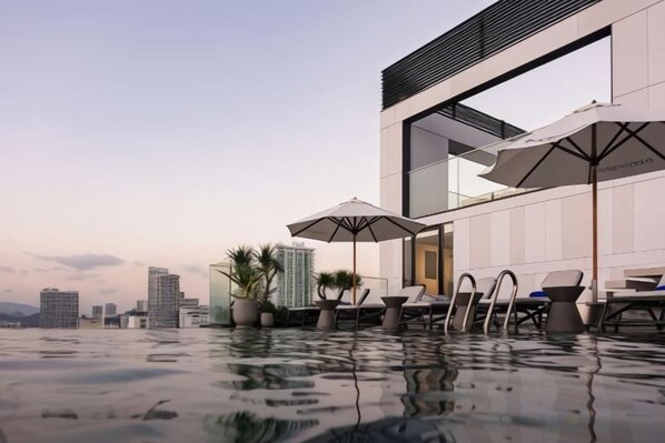 Piscina infinita en la azotea con sillas de descanso y sombrillas, con vistas a un paisaje urbano.