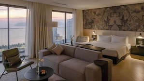 A hotel room with a large bed, a sofa, a small table, and a view of the cityscape through large windows. - December Hotel (Nha Trang)