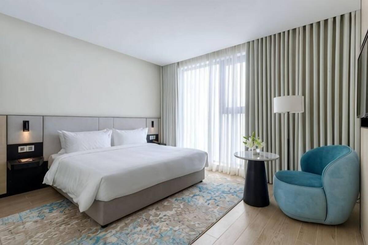 A modern hotel room with a large bed, bedside tables, a round side table, a blue armchair, and a large window with curtains.