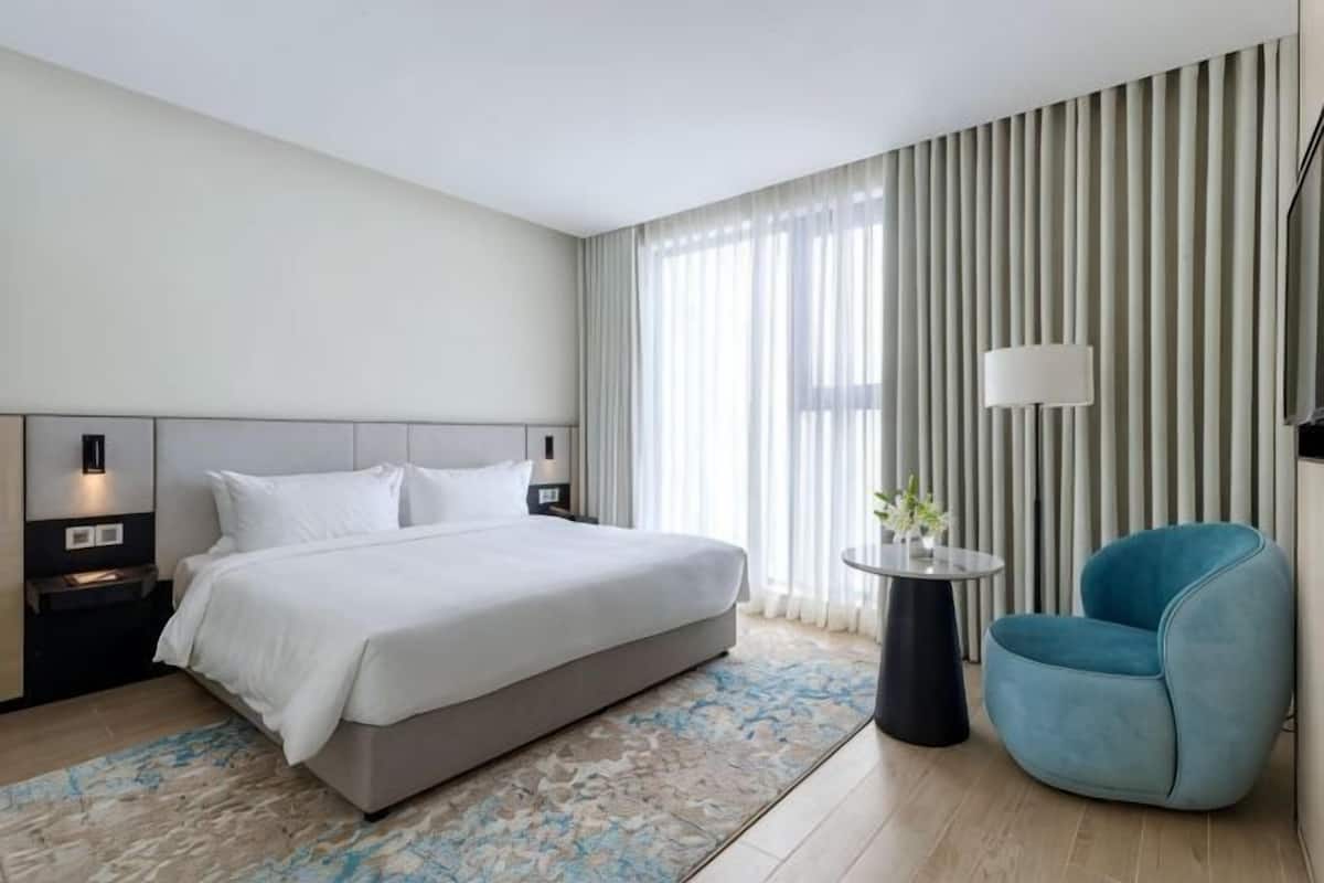 A modern hotel room with a large bed, bedside tables, a round side table, a blue armchair, and a large window with curtains.
