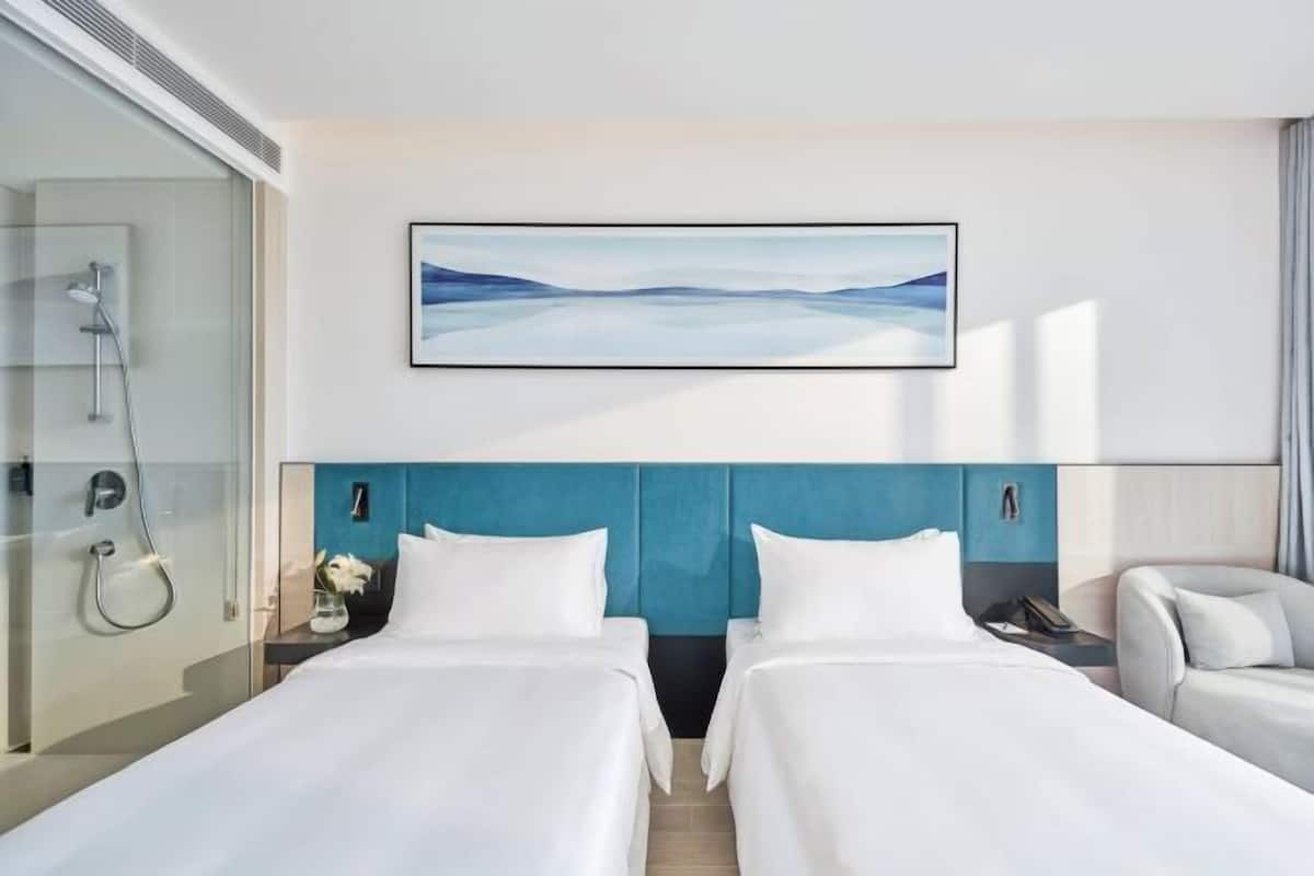 A hotel room with two beds, a large window, a shower area, and a painting on the wall.