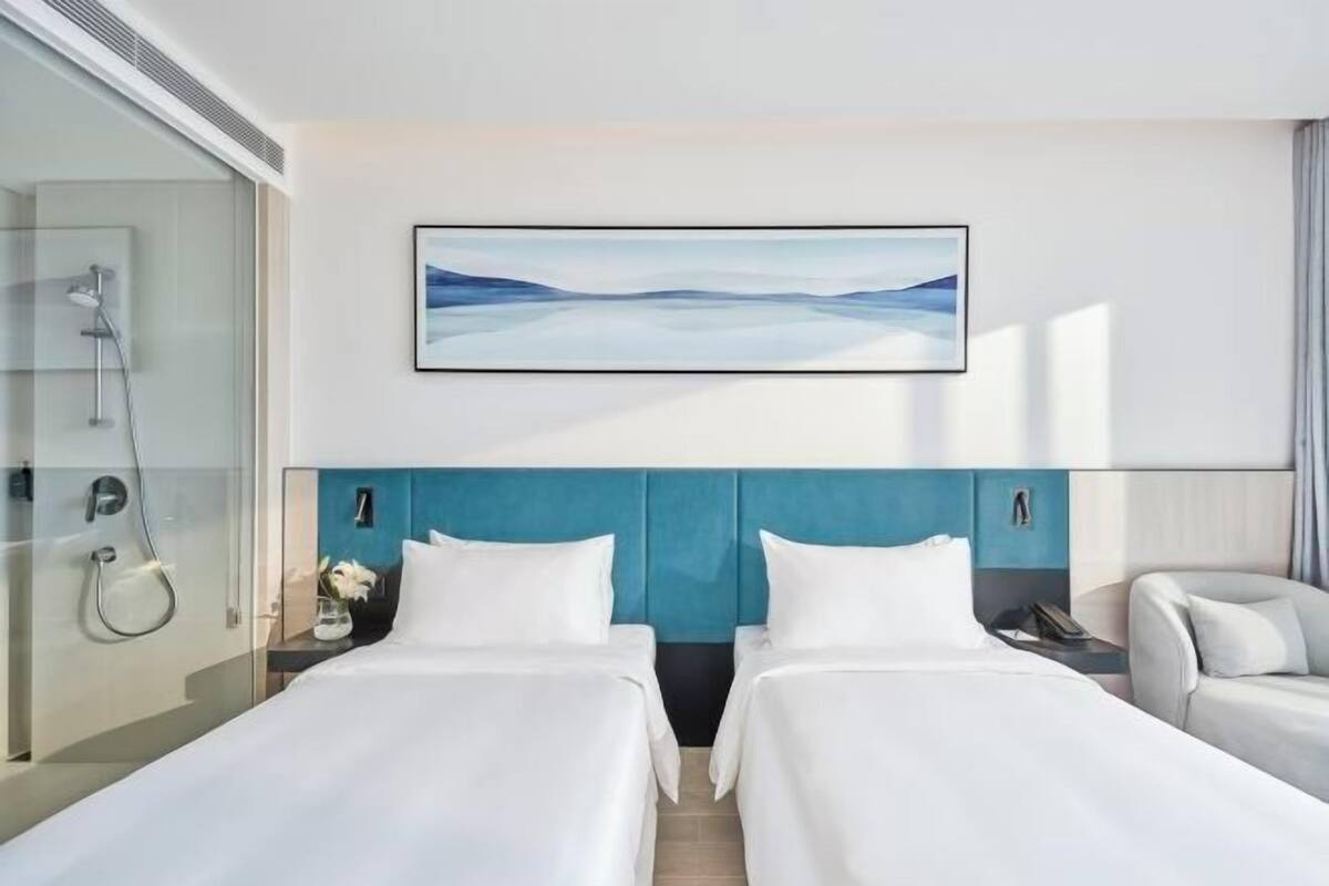 A hotel room with two beds, a shower, a painting on the wall, and a view of the outdoors.