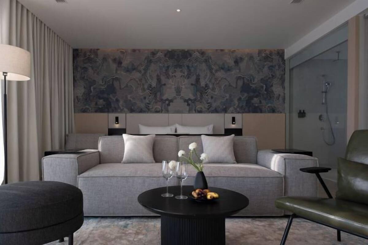 A modern hotel room with a grey sofa, a round black coffee table with a vase and fruit, a green armchair, and a large abstract wall mural.