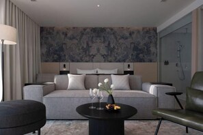 A modern hotel room with a grey sofa, a round black coffee table with a vase and fruit, a green armchair, and a large abstract wall mural. - December Hotel (Nha Trang)