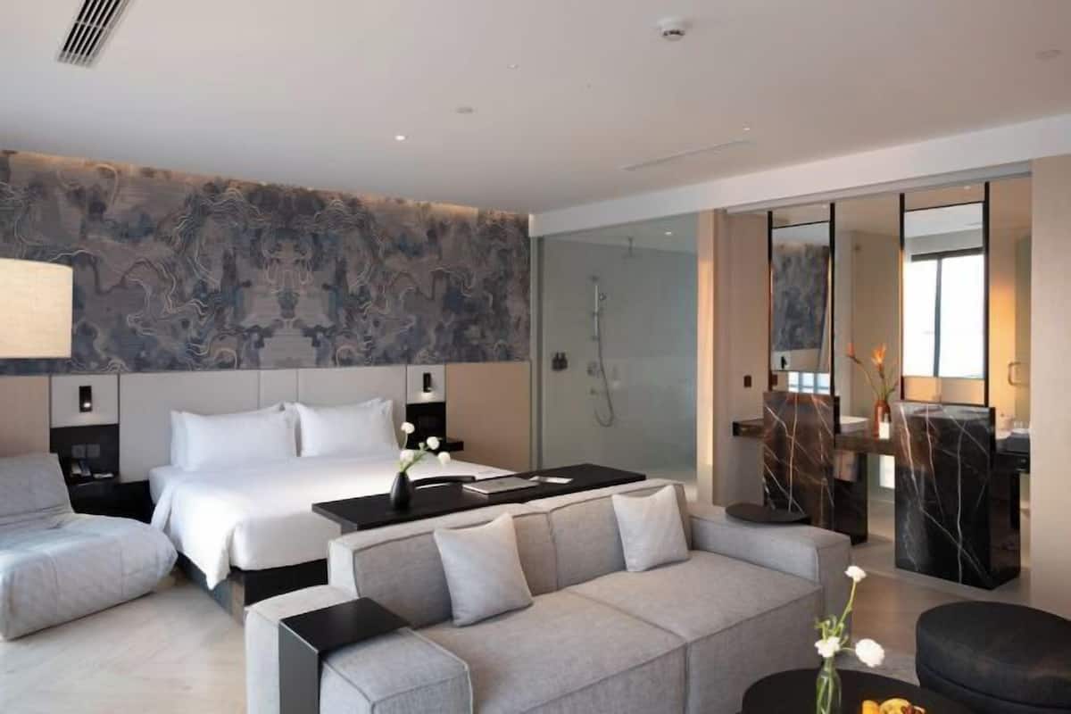 A modern hotel room with a large bed, a sofa, a desk, and a glass shower area.