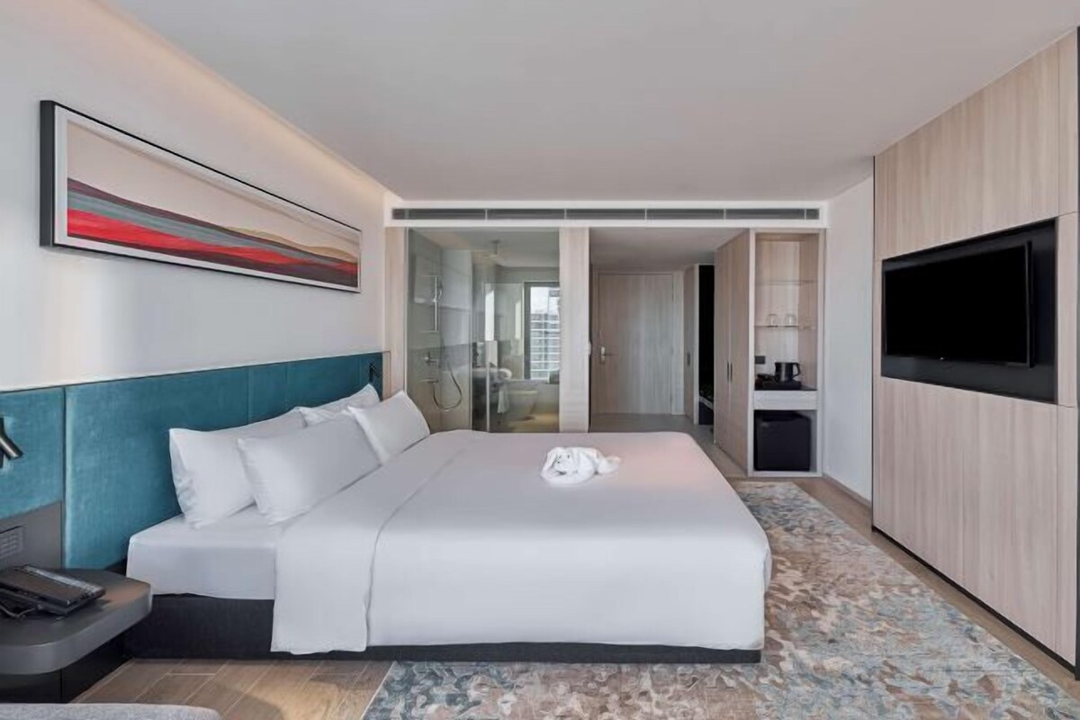 A modern hotel room with a large bed, a flat-screen TV, a desk, and a glass-enclosed bathroom.