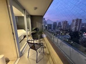 Varanda com guarda-corpo de vidro, cama, mesa e cadeiras.