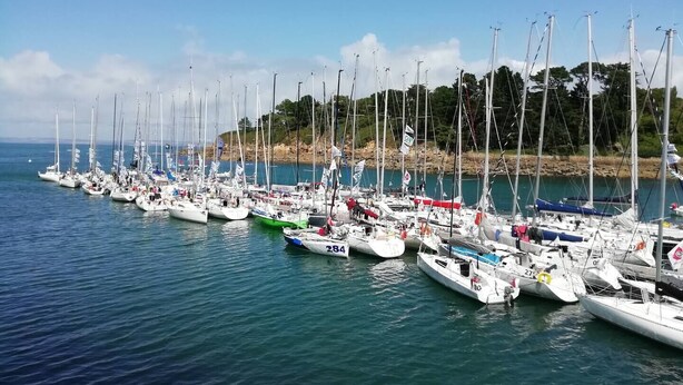 A marina with numerous sailboats docked in calm waters, surrounded by a hilly landscape with trees.