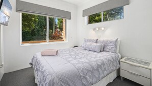 Une chambre avec un grand lit, deux fenêtres équipées de stores, une table de chevet avec une lampe, et une vue sur la verdure à l’extérieur.