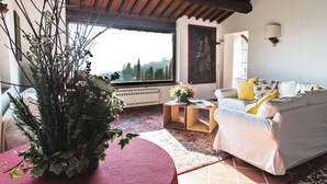 Una sala de estar acogedora con un sofá blanco, una mesa roja y un amplio ventanal con vistas a árboles.