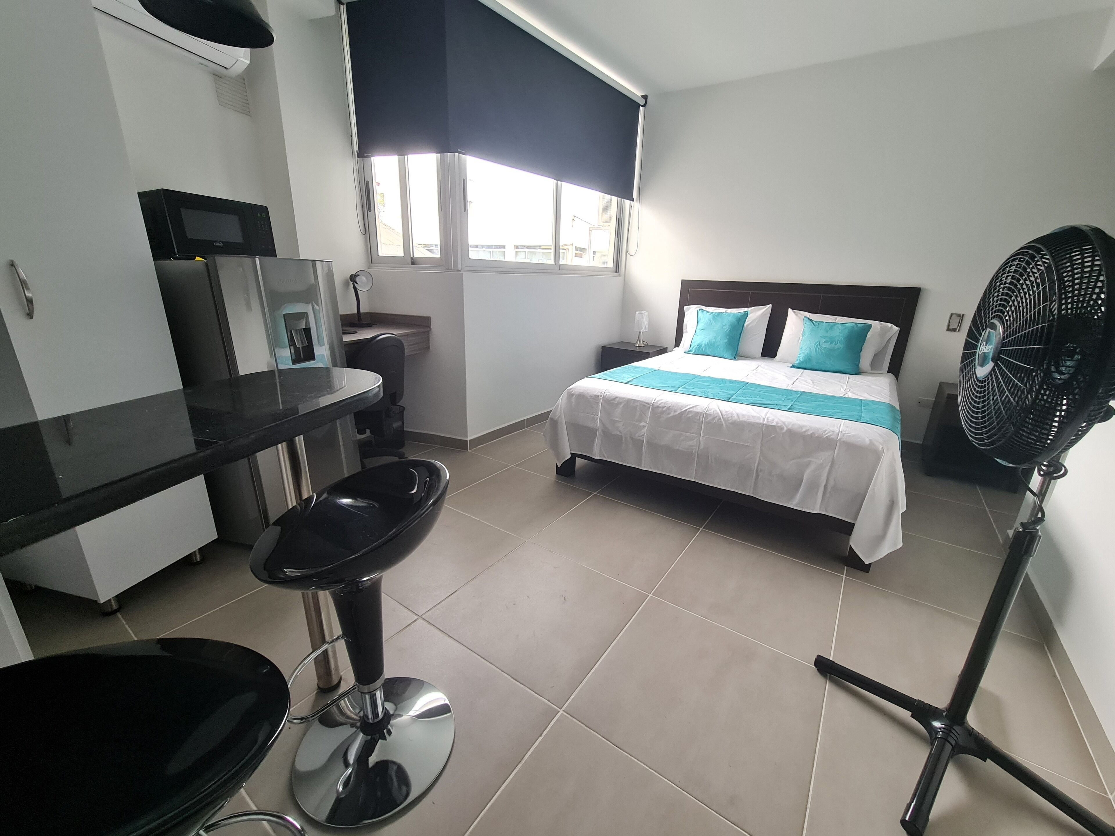 Quarto de hotel com cama, escrivaninha, cadeira, ventilador e janela.