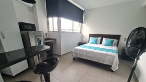 A hotel room with a bed, a desk, a chair, a fan, and a window. - 402 Moderno Aparta-Suite en Versalles Tipo Loft - Cali Tower Suites & Lofts (Cali)