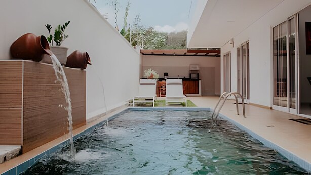 Un espace piscine extérieur moderne, agrémenté d’un élément décoratif aquatique, d’une terrasse en bois et d’un coin cuisine visible au fond.