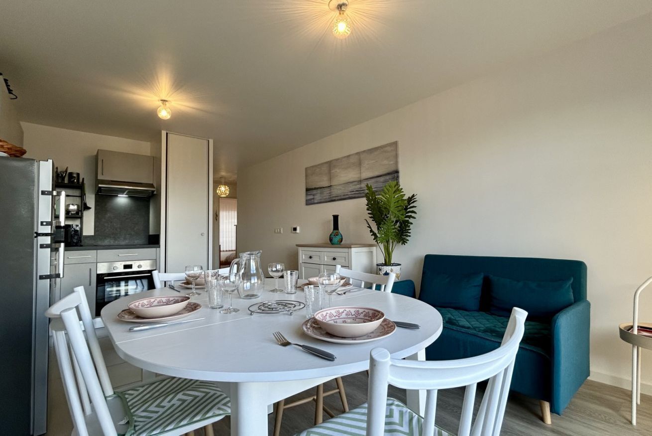 A dining area with a white table set for two, a kitchen in the background, and a teal sofa.