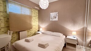 Ein Hotelzimmer mit einem Bett, zwei Nachttischen mit Lampen, einem Sessel, einem Heizkörper und einem Fenster mit Jalousien.