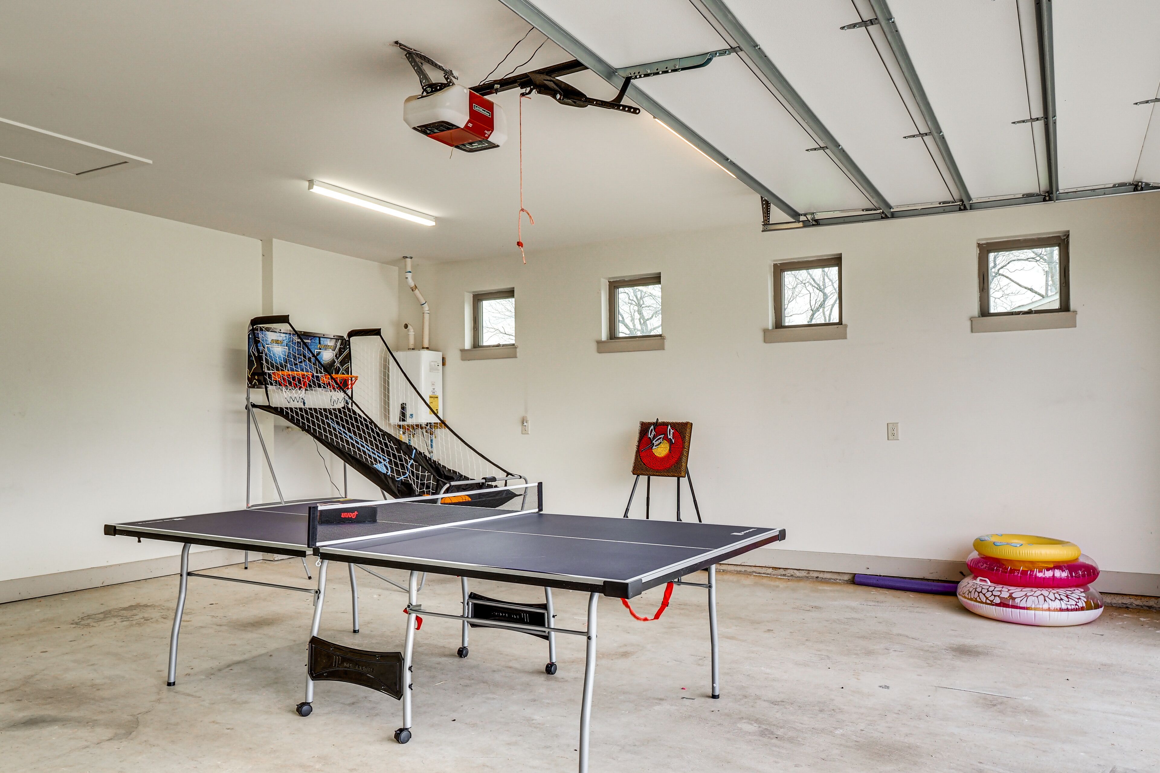 A ping pong table in a room with a ceiling-mounted projector, windows, and a basketball hoop.