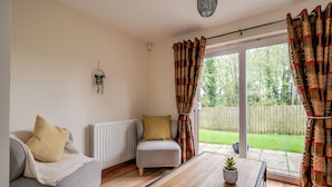 A cozy living room with a sofa, a small table, and a chair. There is a window with curtains, a radiator, and a hanging plant. - Stylish & spacious entire 3+1BR house in Lisburn(near Belfast)with parking  (Lisburn)
