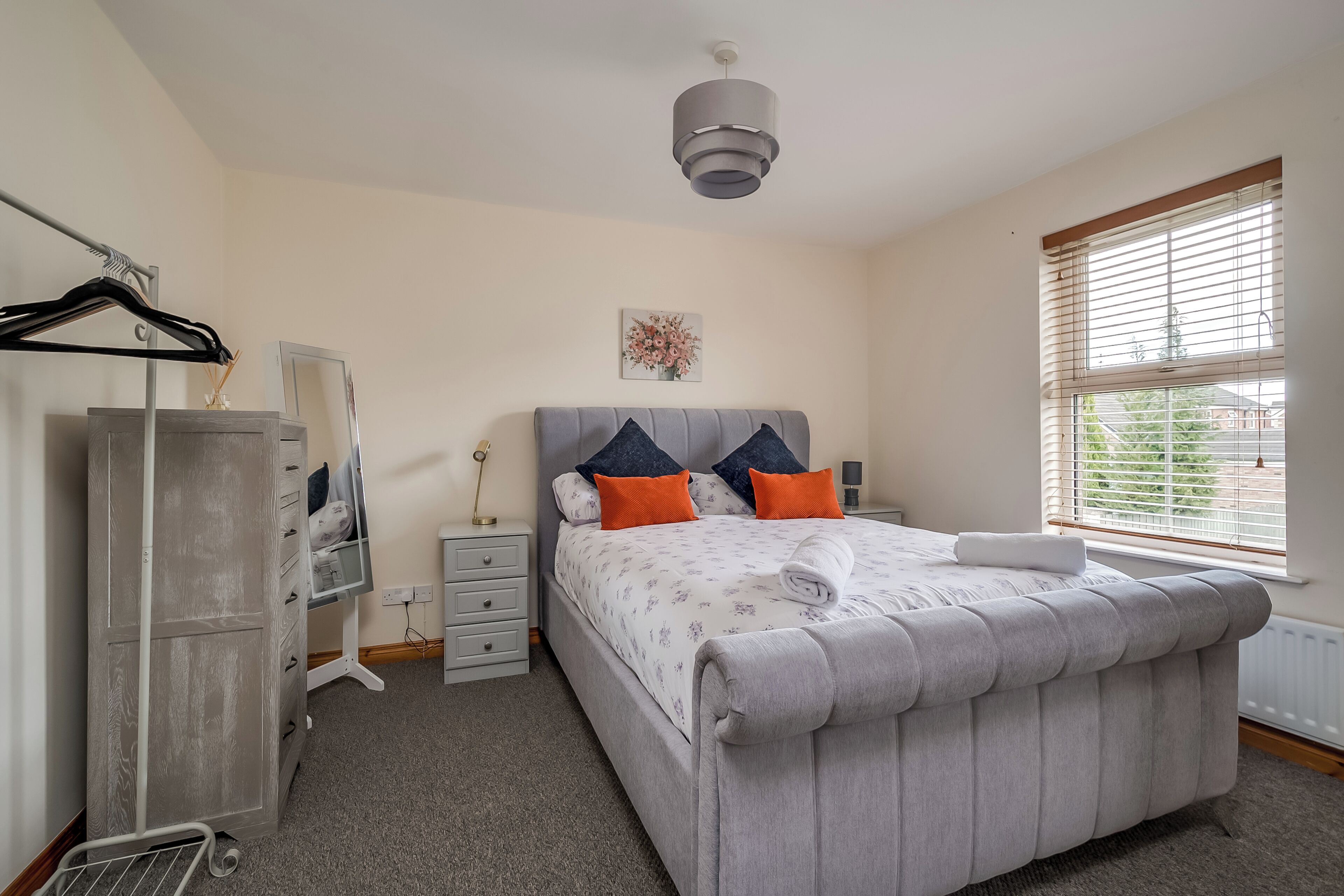 Une chambre avec un lit recouvert d’un tissu gris, deux coussins orange, une serviette blanche, une table de chevet avec une lampe, une armoire et une fenêtre avec des stores.