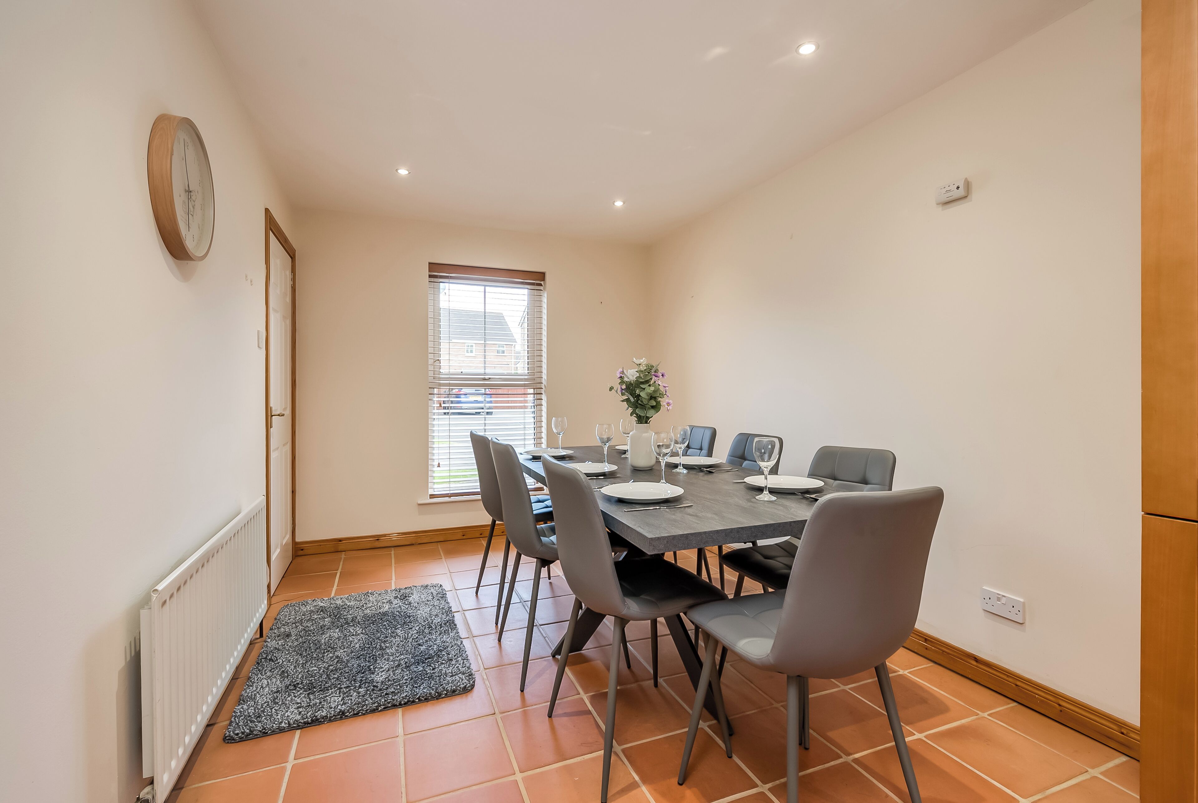 A dining area with a table set for four, grey chairs, a grey rug, a window with blinds, and a wall-mounted clock.
