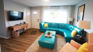 A living room with a teal sectional sofa, a wooden coffee table with a plant, a television mounted on the wall, and a window with sheer curtains. - Charming 3 bedroom retreat (Herrin)