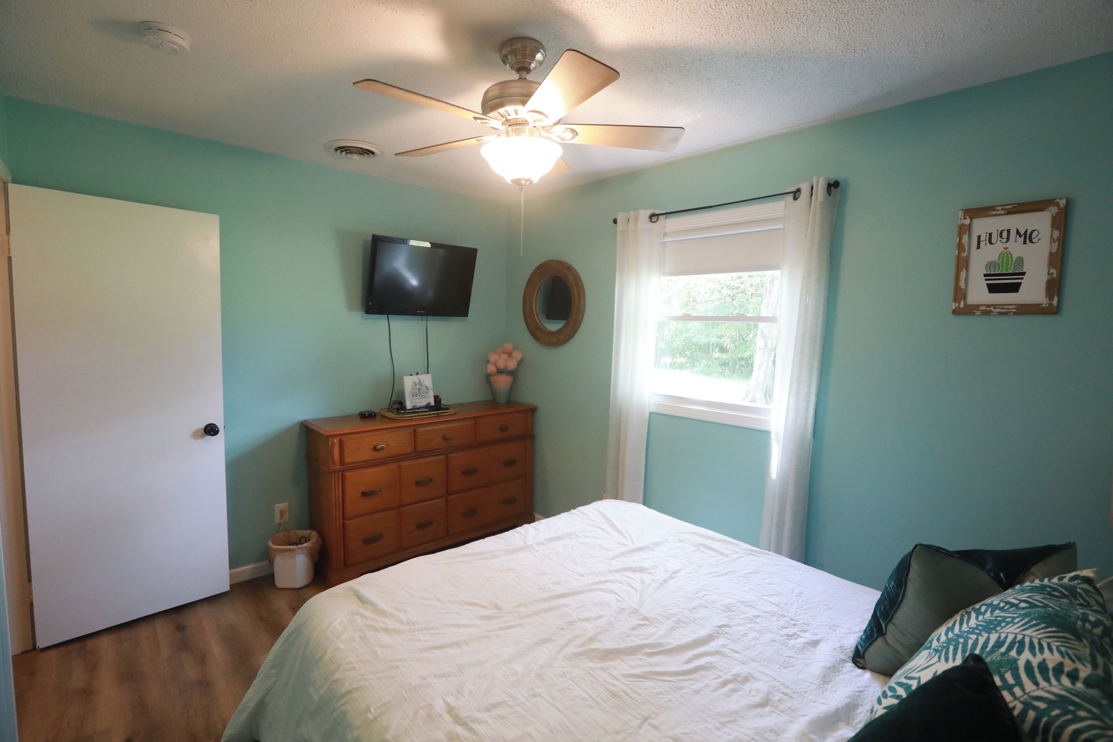 Un dormitorio con cama, ventilador de techo, cómoda, televisor, ventana con cortinas y un adorno en la pared.