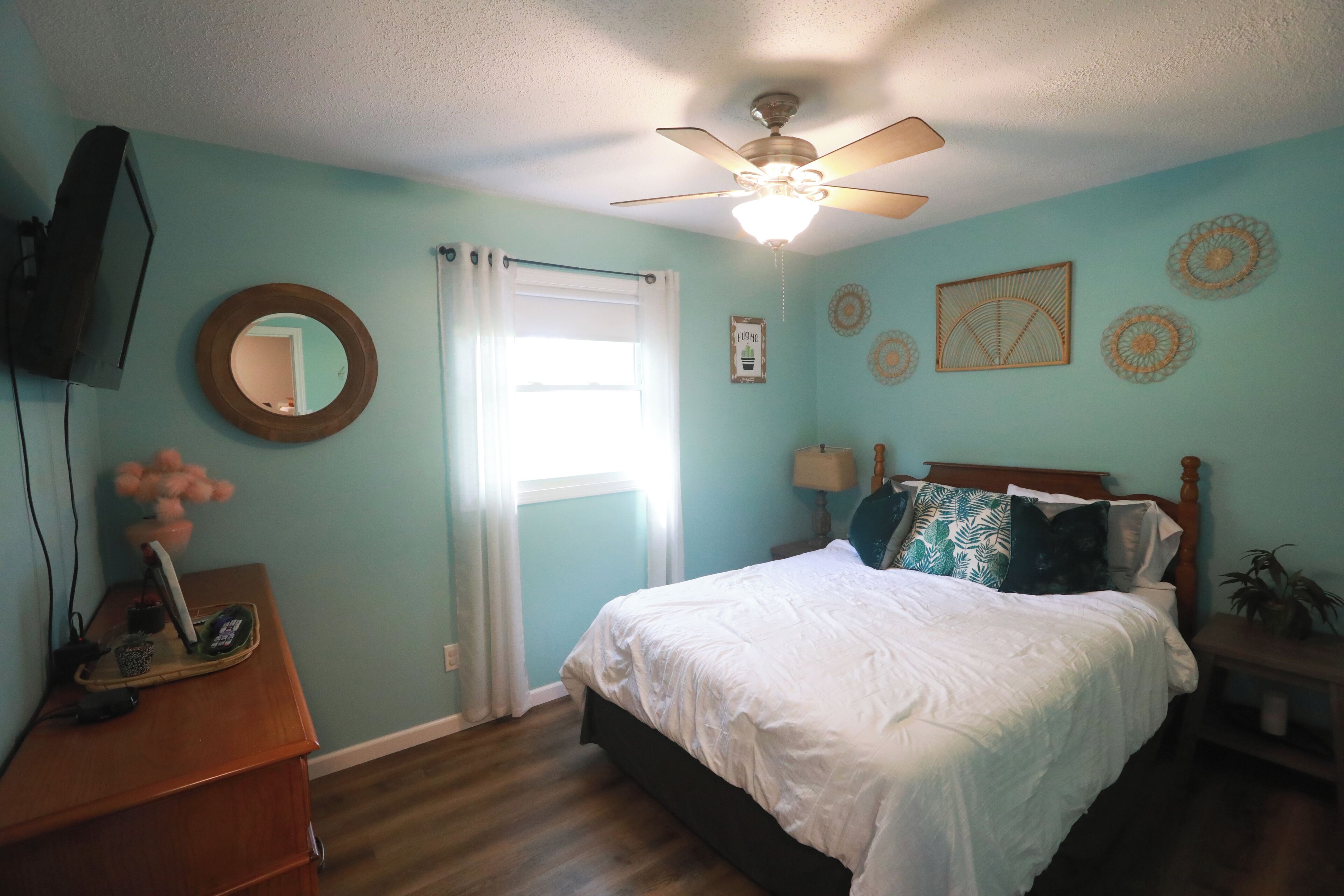 A bedroom with a bed, a ceiling fan, a TV, a dresser, and a window with curtains.