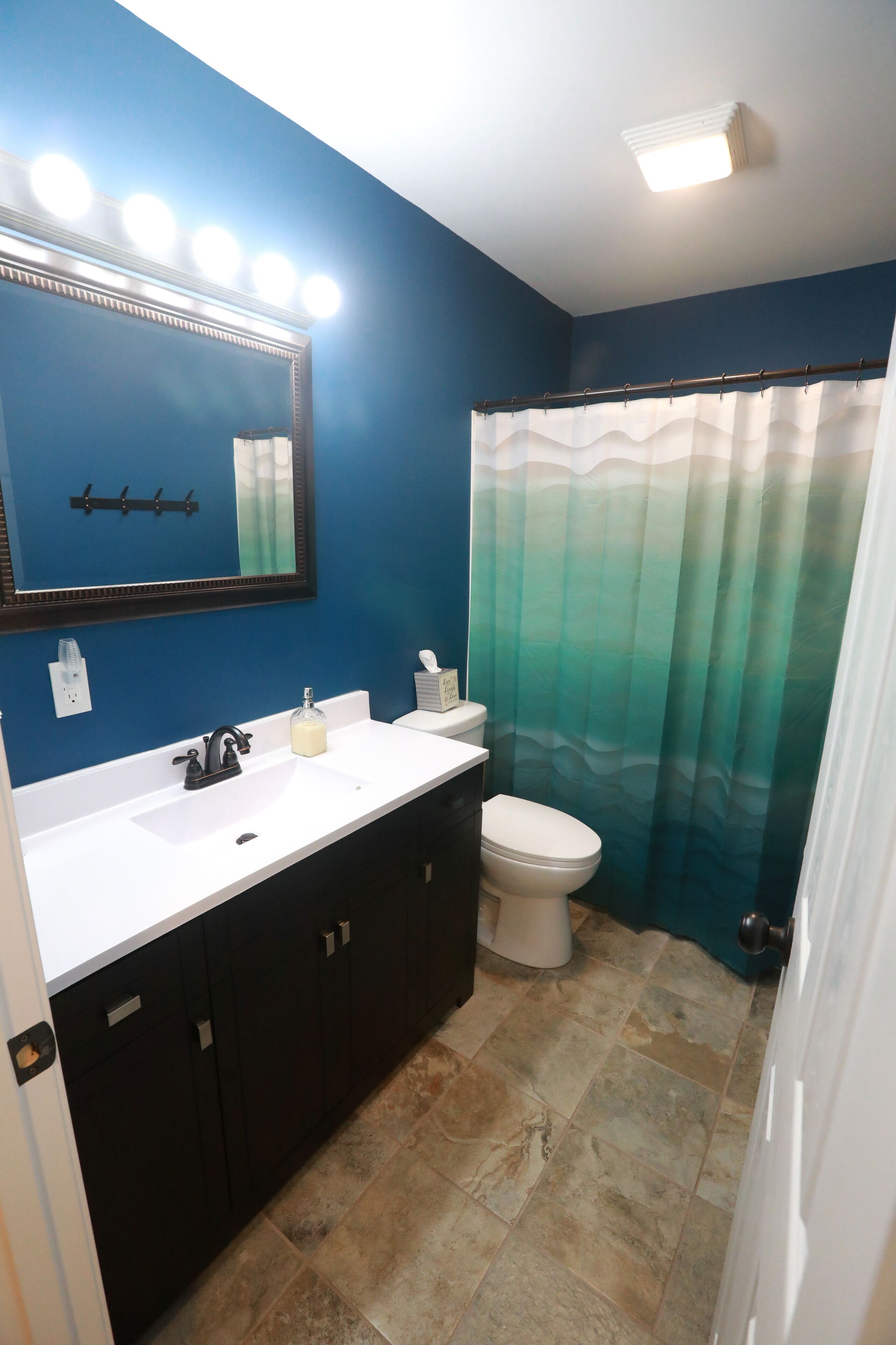 A bathroom with a white sink, dark cabinets, a toilet, a mirror with lights, and a greenish shower curtain.