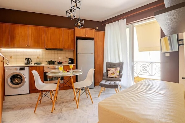 A compact kitchen with wooden cabinets, a washing machine, a dining table with chairs, a gray armchair, and a bed.