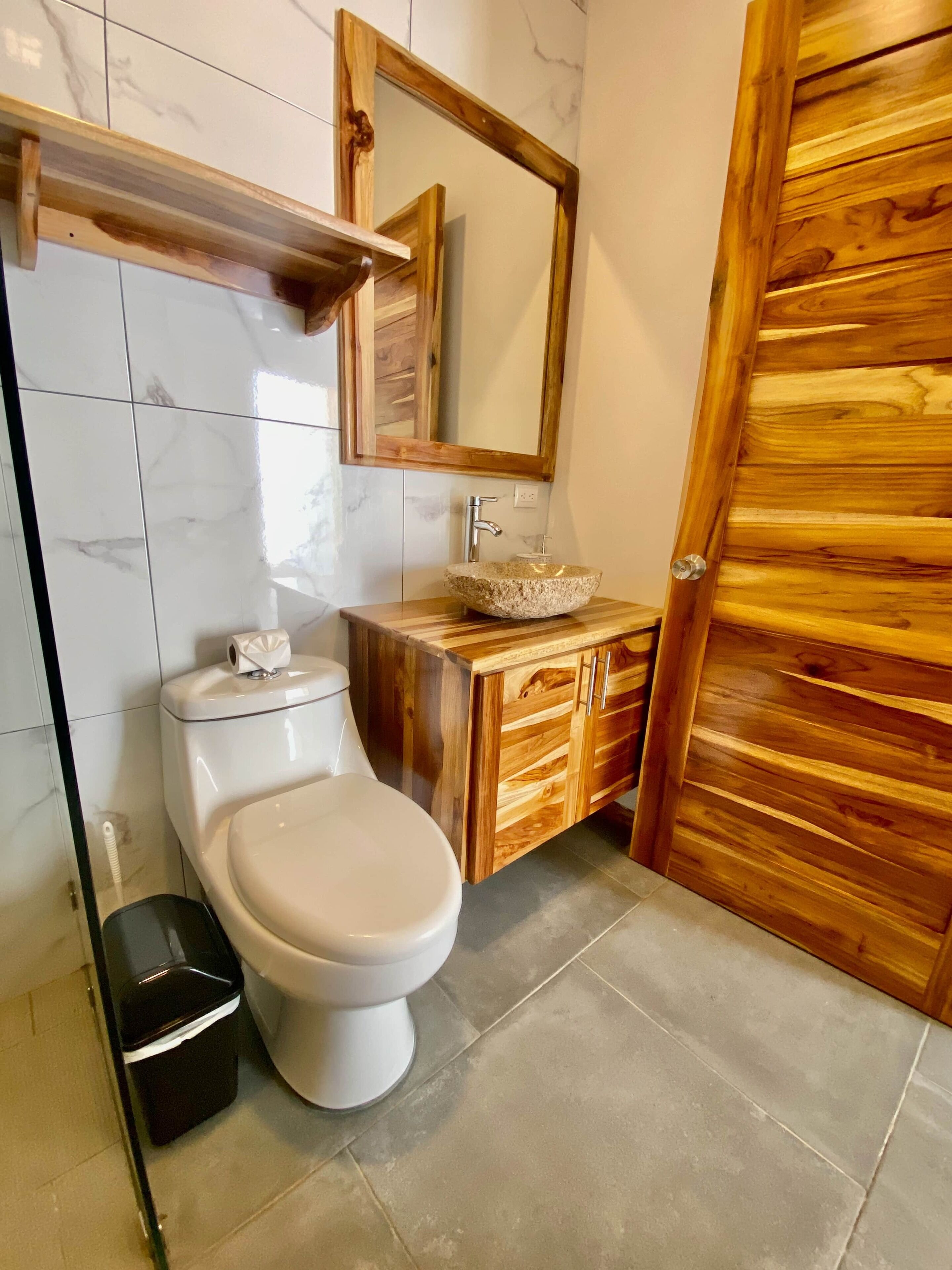 Et badeværelse med et marmortapet gulv, et hvidt toilet, en trækommode med en sten-vask og en trædør.