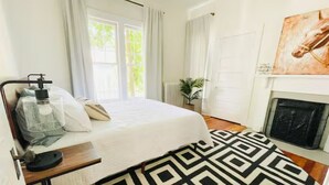 Um quarto com uma cama, uma mesinha de cabeceira com abajur, um tapete estampado, uma lareira e um quadro na parede.