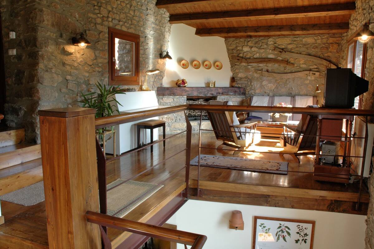 A rustic interior with a stone wall, wooden beams, a fireplace, and a wooden staircase.