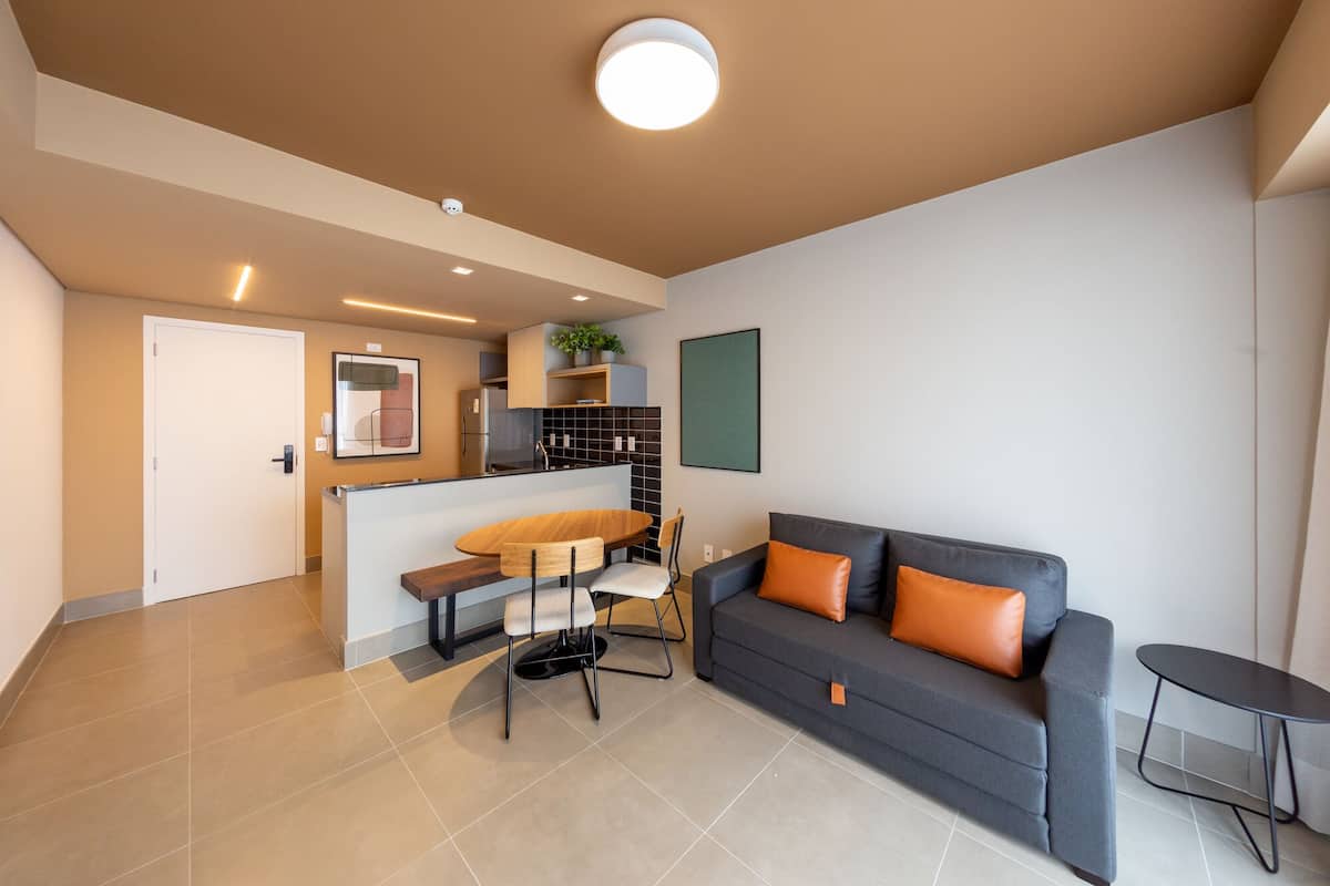Uma sala de estar moderna com um sofá cinzento, uma mesa redonda pequena e uma área de cozinha com geladeira e uma planta.