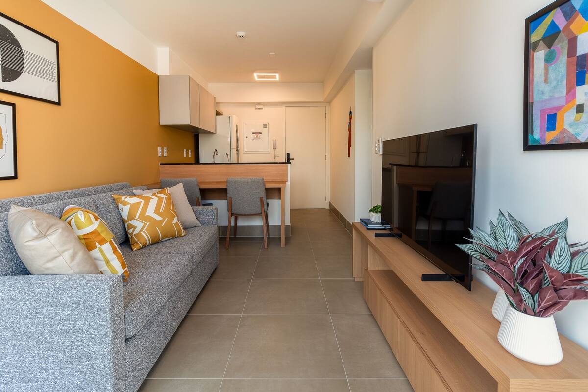 Uma sala de estar moderna com um sofá cinzento, um móvel para televisão de madeira e uma área de refeições ao fundo.