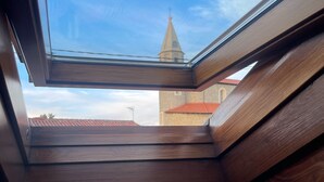 Une lucarne offrant une vue sur le clocher d’une église et sur des murs recouverts de tuiles.