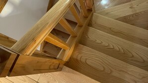 Un escalier en bois, doté d’une rampe et d’un élément décoratif en forme de boule, mène à un palier avec un mur blanc et un plancher en bois.