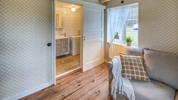 Een gezellige kamer met een houten vloer, een bank met een ruitkussen, een raam met een plant en een badkamer met een witte deur en betegelde muren.