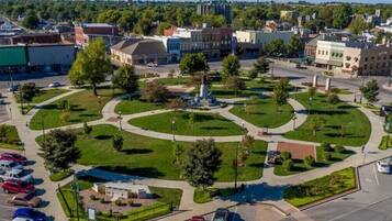 Una piazza pubblica con alberi e aiuole, dominata da una fontana centrale, circondata da edifici e un'area di parcheggio.