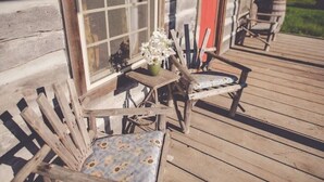 A wooden deck with two chairs and a small table, a window with a view of greenery, and a red door. - Audrey's Abode - Restored log cabin, country setting, minutes from Jacksonville
 (Jacksonville)