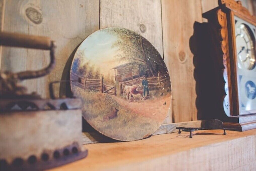 A rustic wooden wall with a round mirror reflecting a farm scene, a vintage clock, and a small metal object on a shelf.