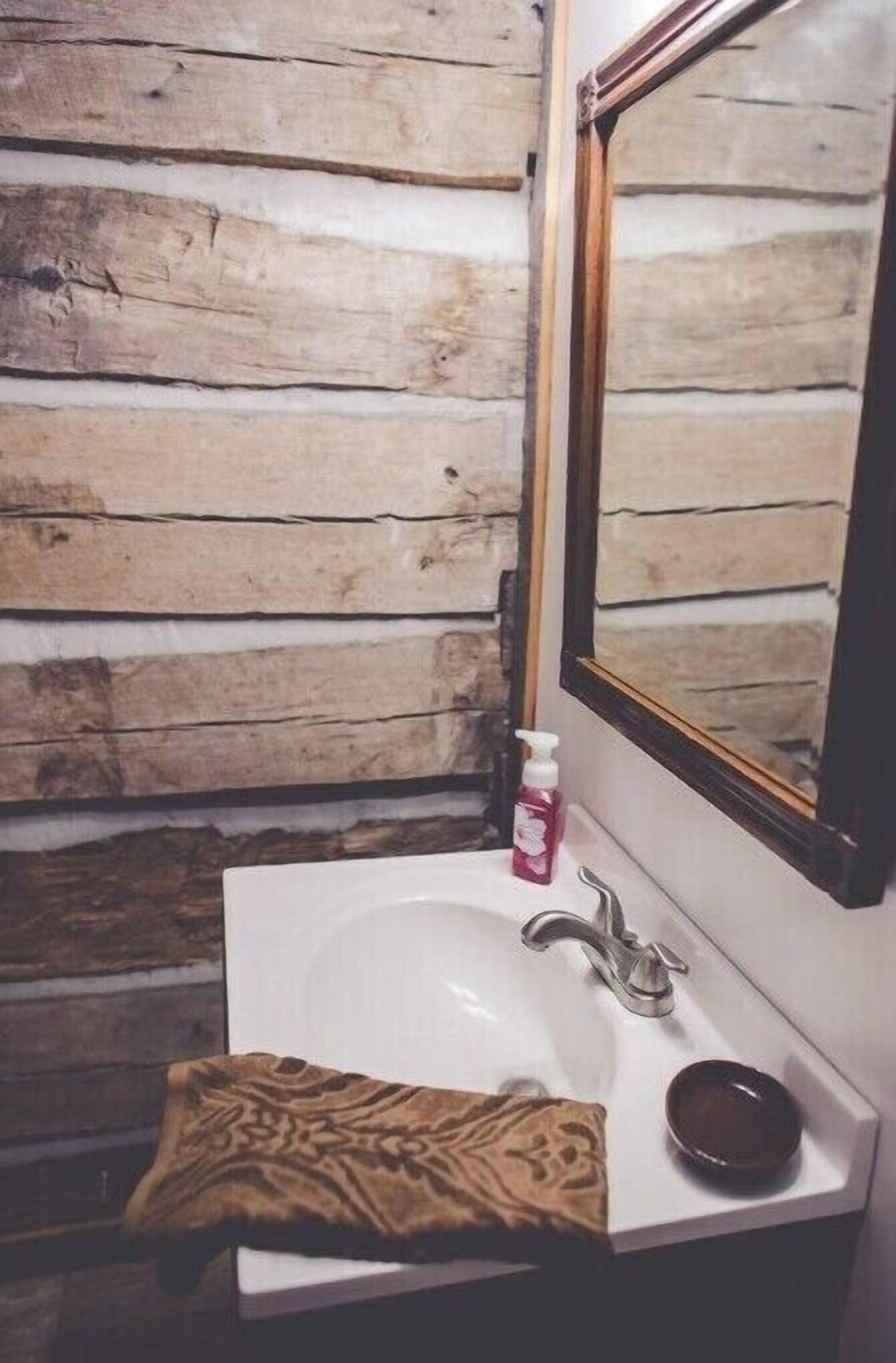 Un baño con pared de madera, un lavamanos blanco, un espejo, una toalla y un dispensador de jabón.