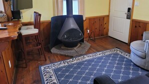 A cozy living room with a wood-burning stove, a television, a dining area with chairs, and a patterned rug. - Hillcrest Suites & Cabins (Big Bear Lake)