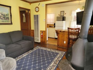 Uma sala de estar com sofá, tapete estampado, lareira e área de cozinha visível por uma porta aberta.
