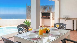 Une table à manger dressée avec des assiettes, des verres et des fruits, donnant sur une piscine et offrant une vue sur l’océan.