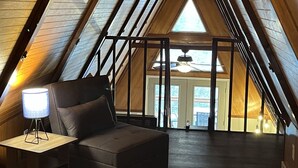 Una camera da letto mansardata accogliente, con soffitto inclinato, un divano confortevole, un piccolo tavolo con lampada e vista all'esterno tramite un'ampia finestra.
