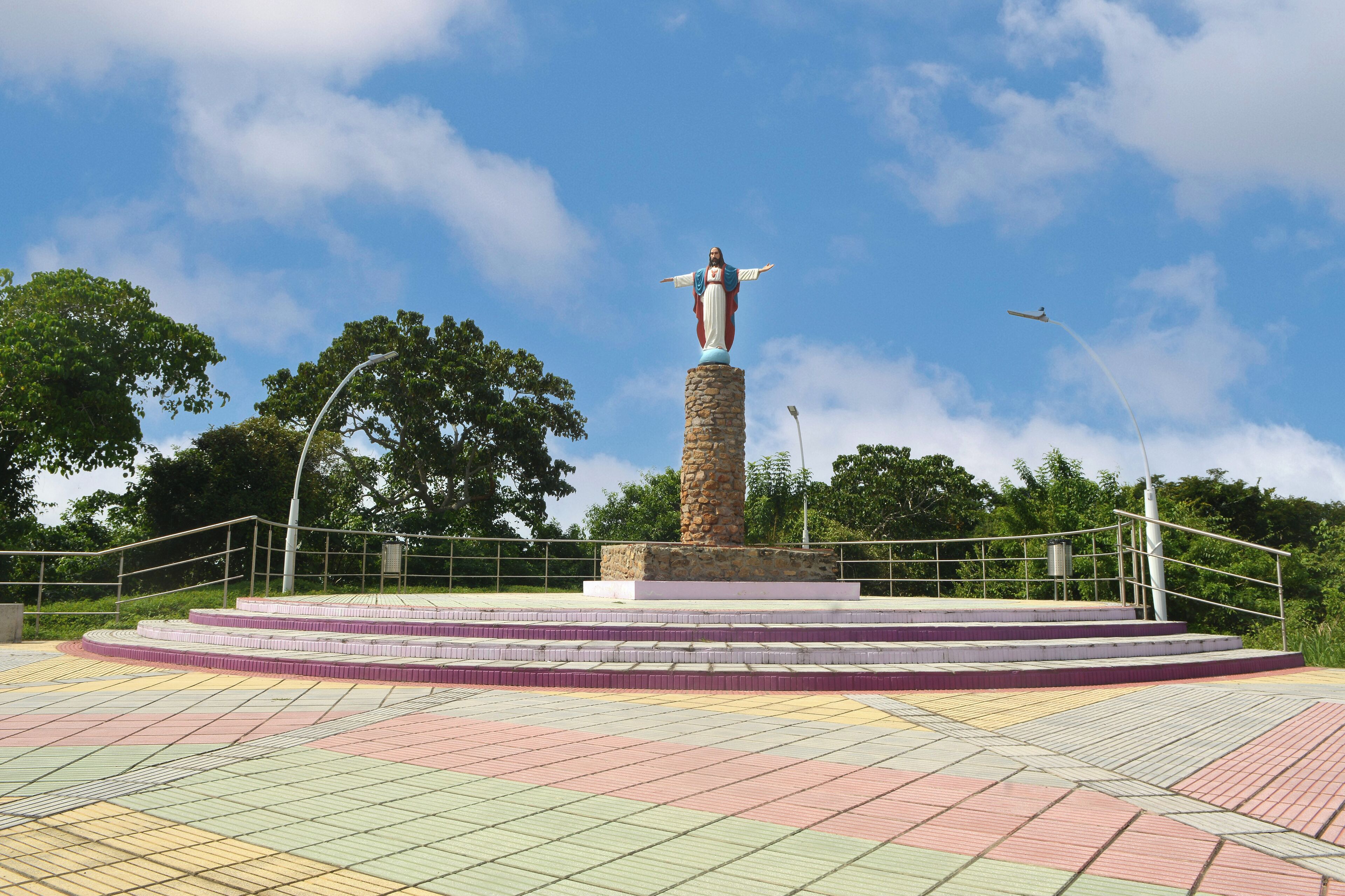Une statue sur une plateforme circulaire accessible par des marches, entourée d’une balustrade et avec des arbres en arrière-plan.