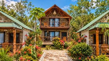 A two-story stone building with a balcony, surrounded by lush greenery and flowering plants.